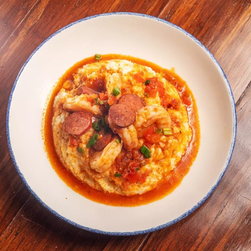 Shrimp and grits with sliced sausage, tomatoes, and green onions in a white bowl.