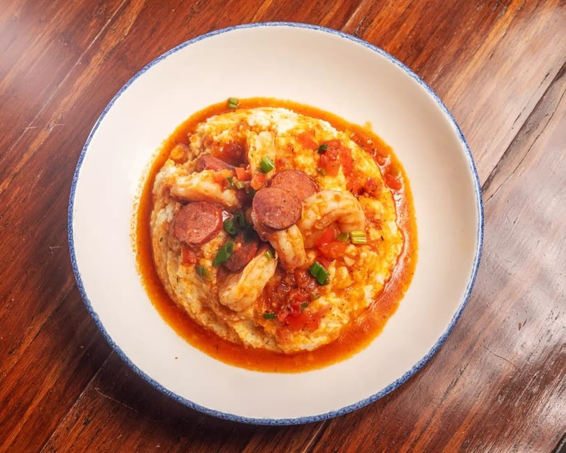 Shrimp and grits with sliced sausage, tomatoes, and green onions in a white bowl.