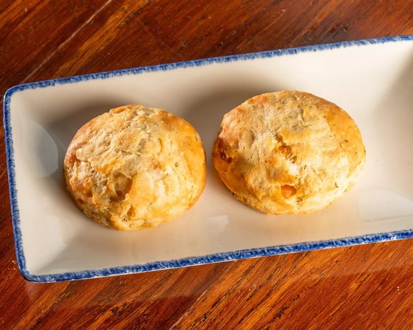 Two golden-brown biscuits with herbs on a white rectangular plate with a blue rim, on a wooden table.