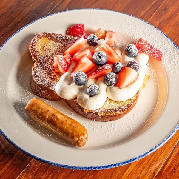 French toast topped with whipped cream, strawberries, blueberries, powdered sugar, and a sausage link.