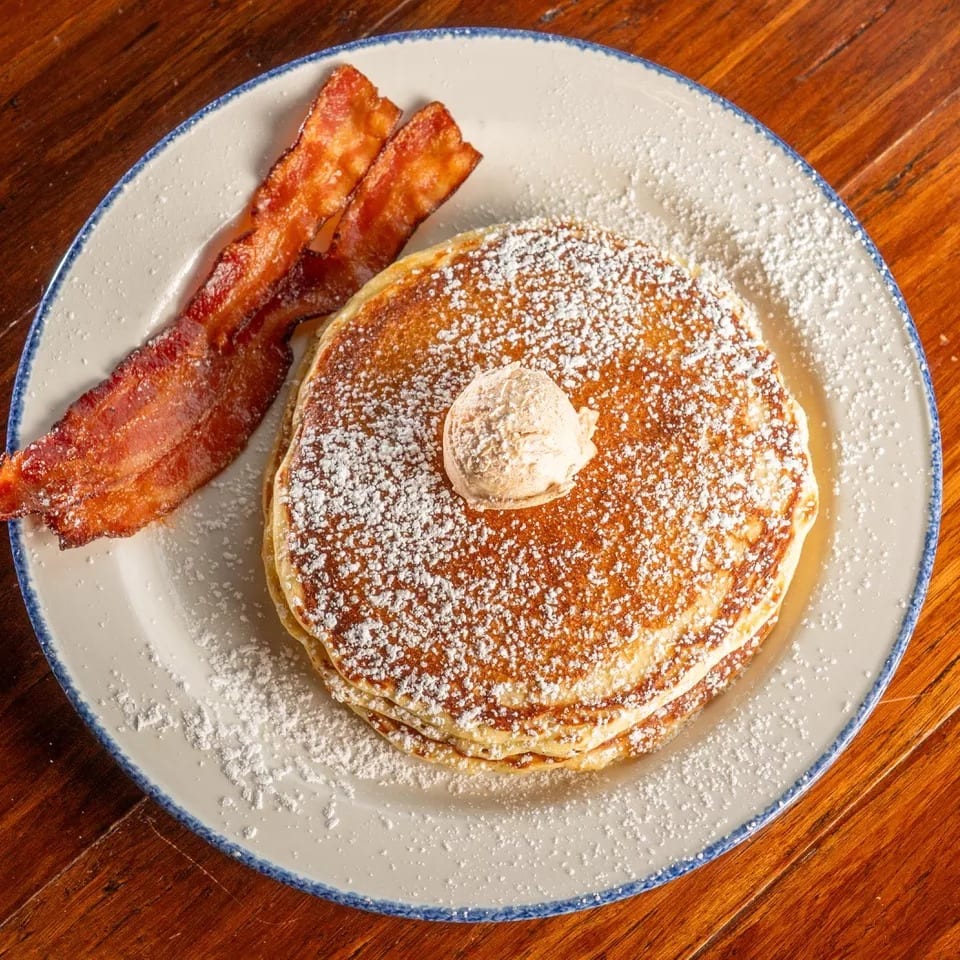 Stack of pancakes with butter and powdered sugar, served with two strips of bacon.