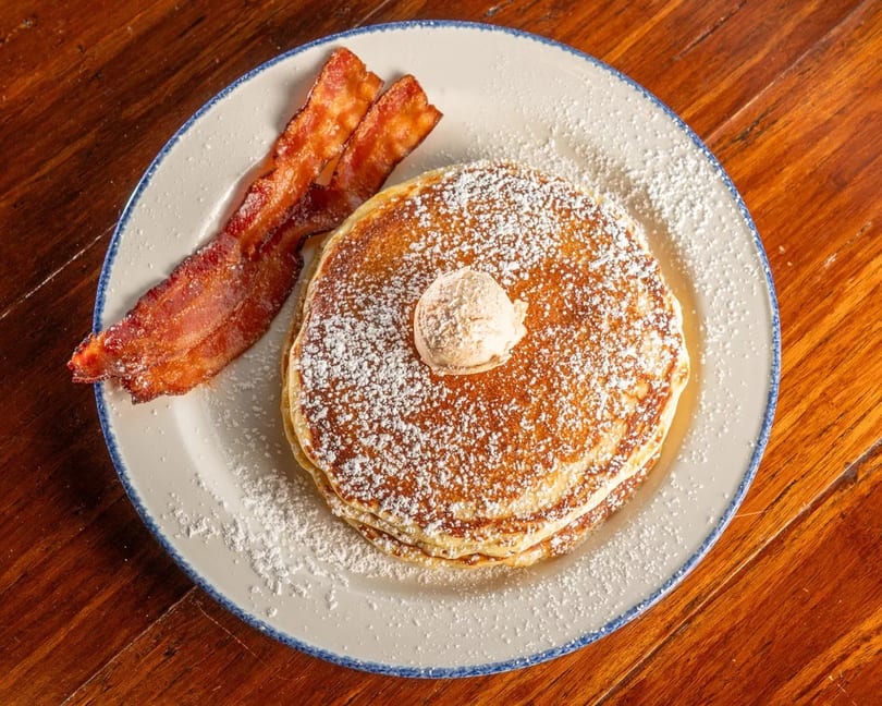 Stack of pancakes with butter and powdered sugar, served with two strips of bacon.